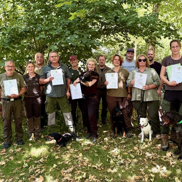 Eine Gruppe Menschen steht im Freien und hält Urkunden in den Händen. Um sie herum sind einige Hunde zu sehen. Im Hintergrund wachsen grüne Bäume.