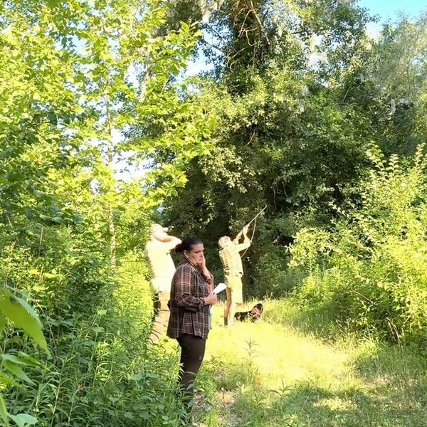Personen stehen in einem dichten, grünen Wald. Eine Person notiert etwas, während andere in die Baumkronen blicken. Sonnenlicht scheint durch das Blätterdach.